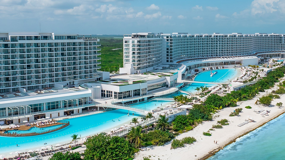 Aerial view of a luxury beachfront resort with white towers, turquoise pools, palm trees, and a sandy beach beside calm ocean.