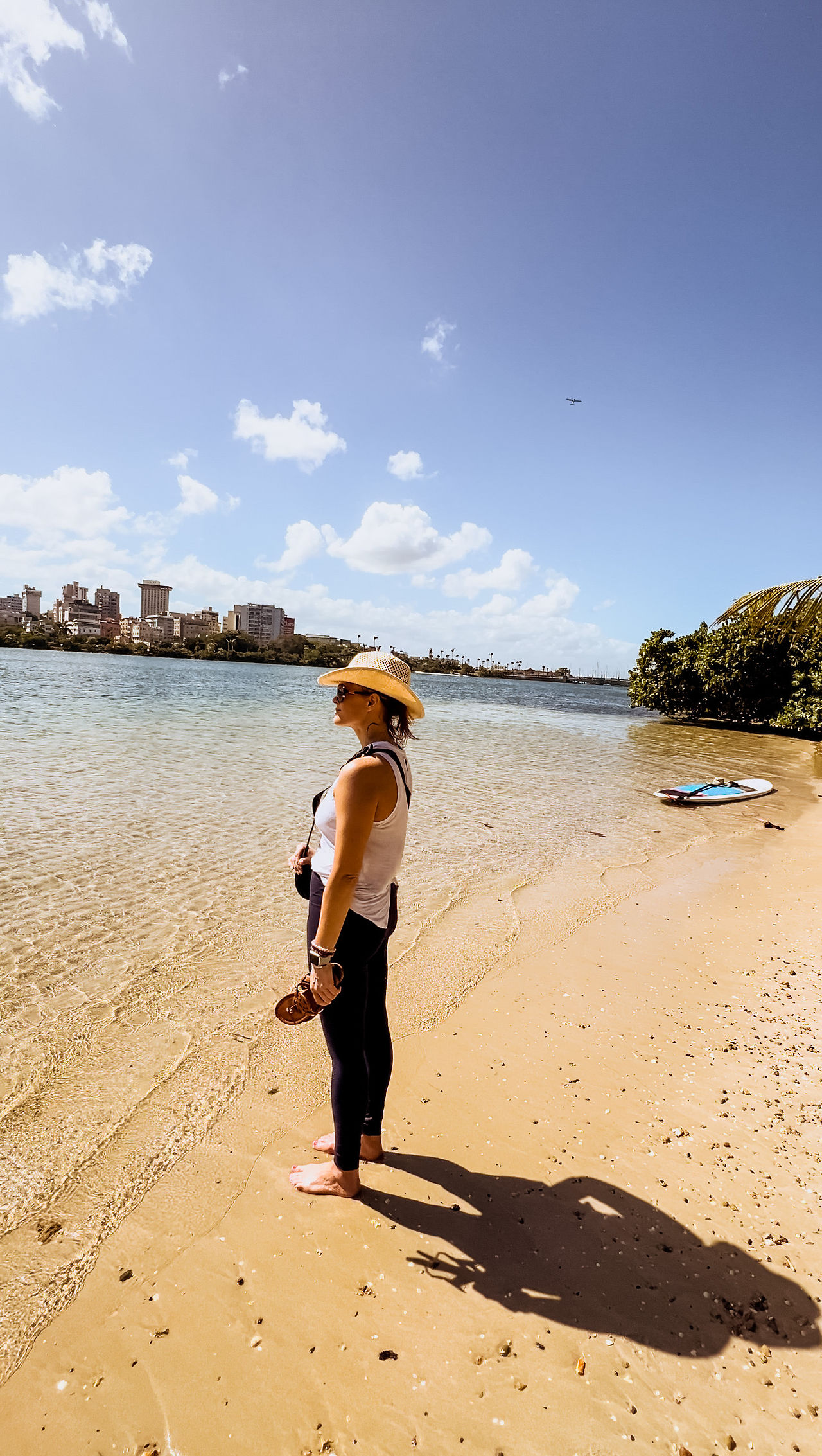 A girl in a har stands barefoot on the beach, holding sandals, gazing at water