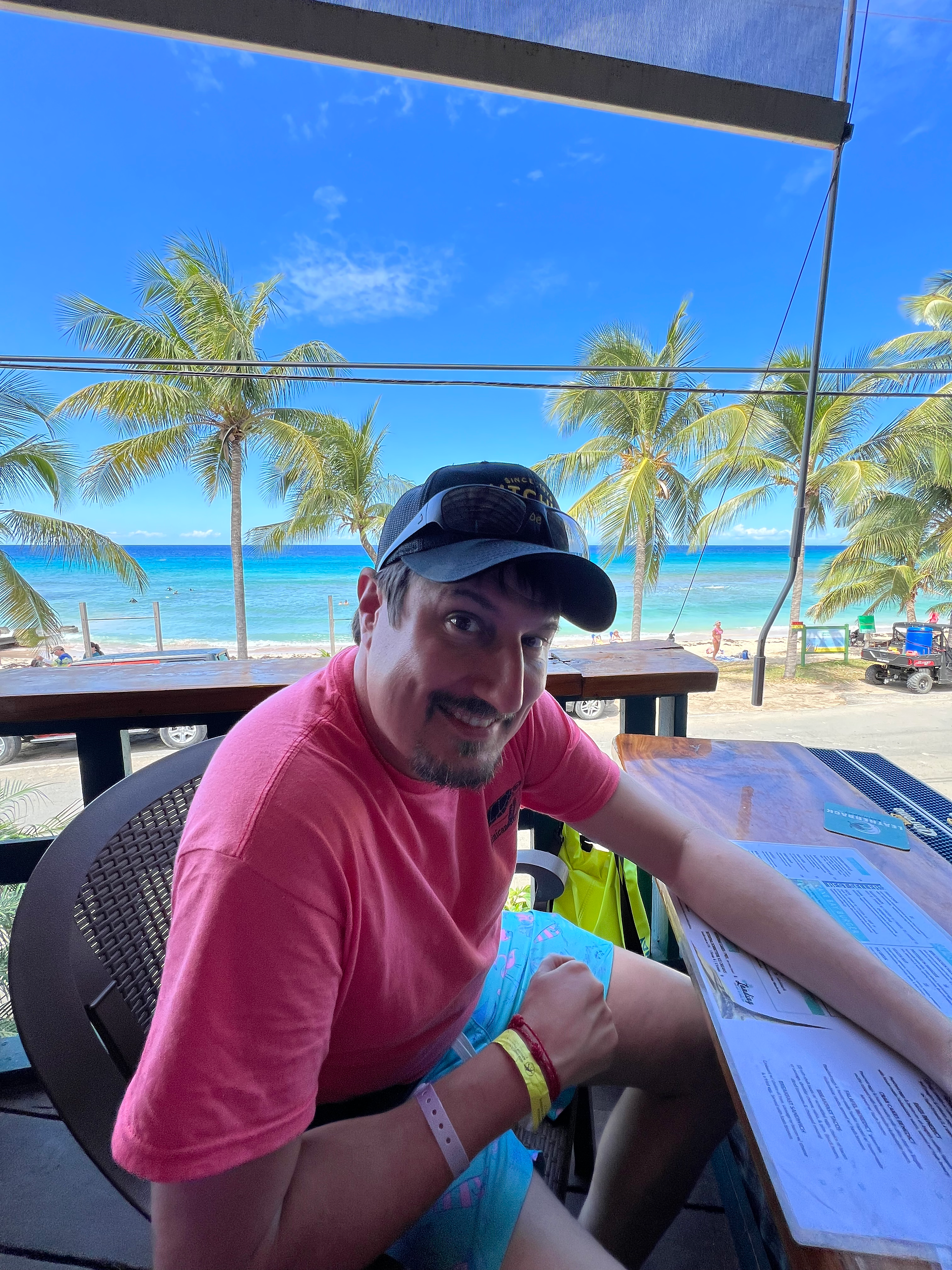 A man sitting an a wooden bar, with beautiful beach and palm trees in the background
