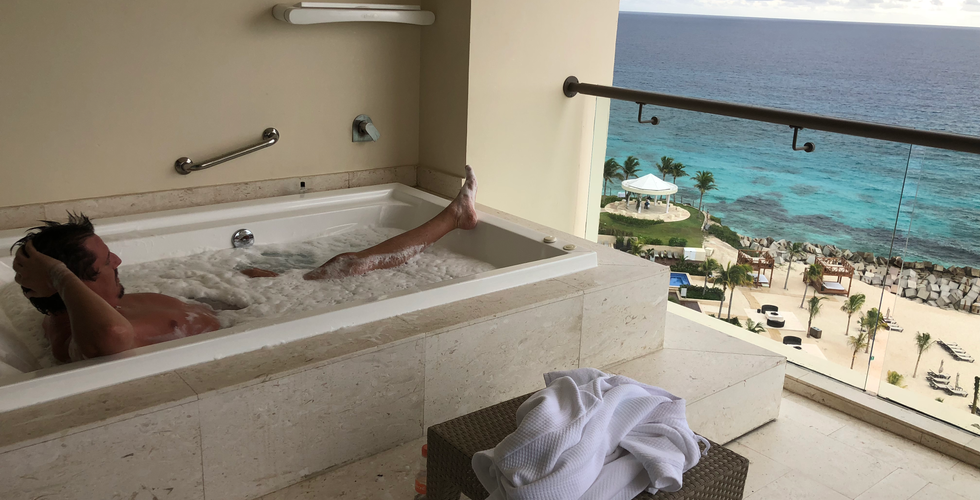 A man relaxing in a hot tub with bubbles on a balcony overlooking the blue ocean at Hyatt Ziva from the Turquoize Tower.
