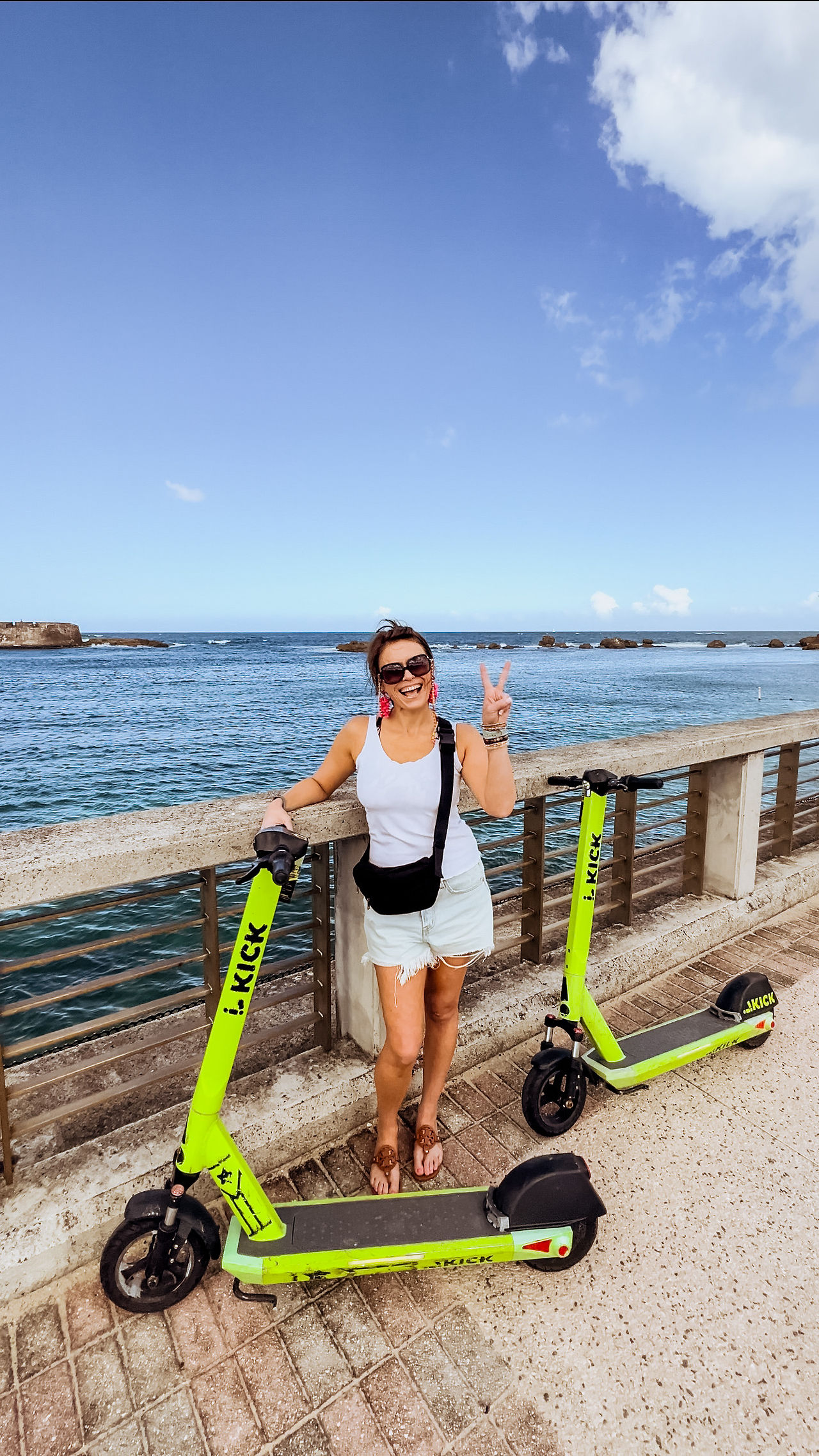 A girl in white tank top stands on a bridge giving the peace sign, standing with a Lime scooter