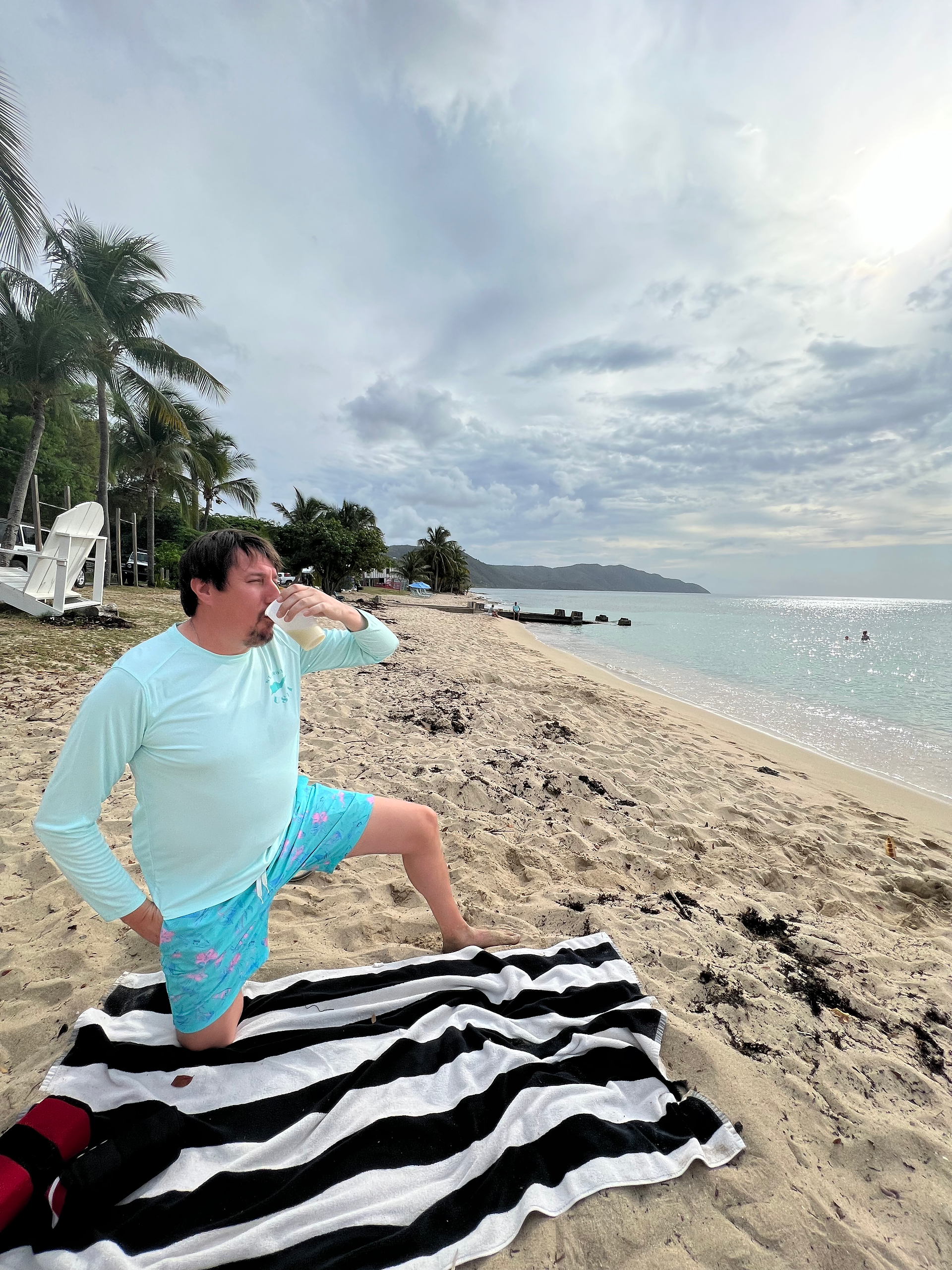 A man dressed in blue swim trunks and shirt kneels on a black and white striped towel on a beach in cane bay