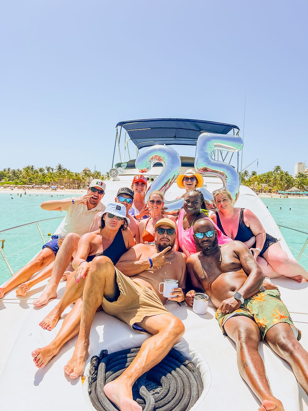 Smiling group lounges on a yacht with silver 25 balloons, drinks, and tropical beach backdrop under a bright blue sky