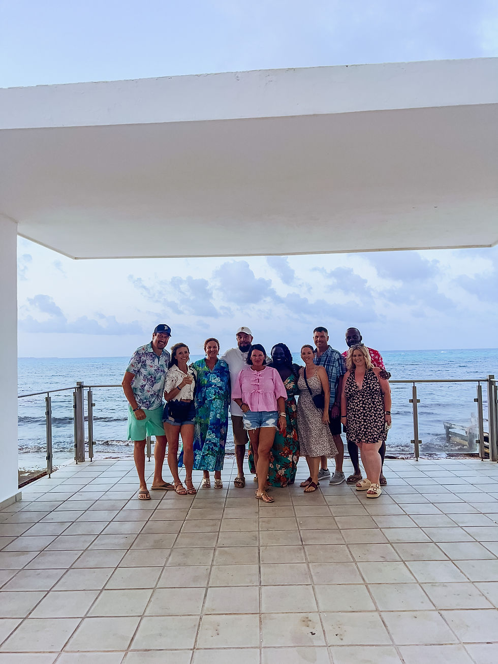 Nine smiling adults pose on a seaside patio under a white canopy, with ocean and cloudy sky behind them.