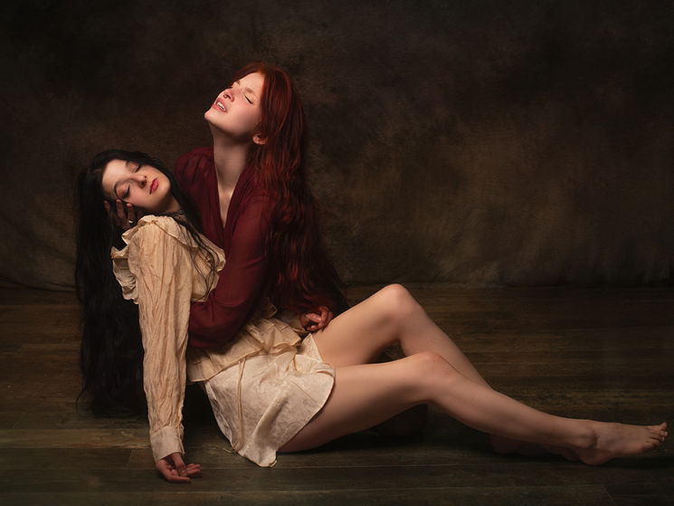 Deux femmes assises au sol, étroitement enlacées. L’une soutient le corps relâché de l’autre, dont la tête repose contre elle, les yeux clos. La scène, éclairée en clair-obscur sur fond sombre, évoque la fragilité, la dépendance et l’effacement progressif de soi dans l’autre.