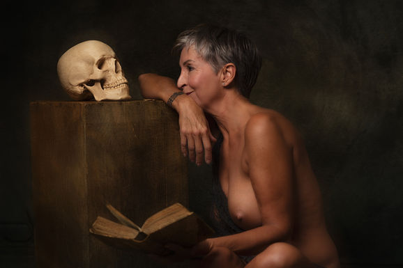 Photographie fine-art montrant une femme mature posant nue en lisant un livre ancien, le regard tourné vers un crâne posé sur un bloc en bois, dans une ambiance intime et symbolique.
