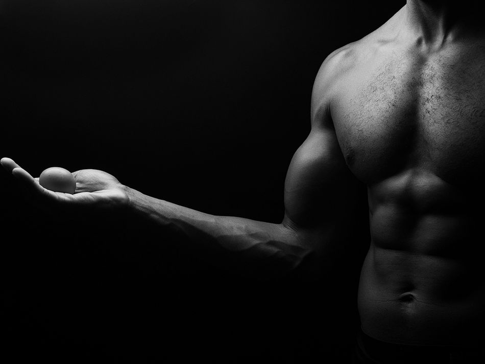 Photographie en noir et blanc montrant le torse d’un corps masculin partiellement éclairé, le bras tendu vers l’avant tenant un objet rond dans la paume de la main. Le fond est entièrement noir, évoquant la force, l’attente et la responsabilité.