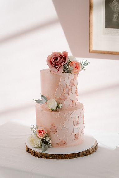 A two-tiered wedding cake with pink textured icing and floral decoration in white and various shades of pink sits on a round wooden board.