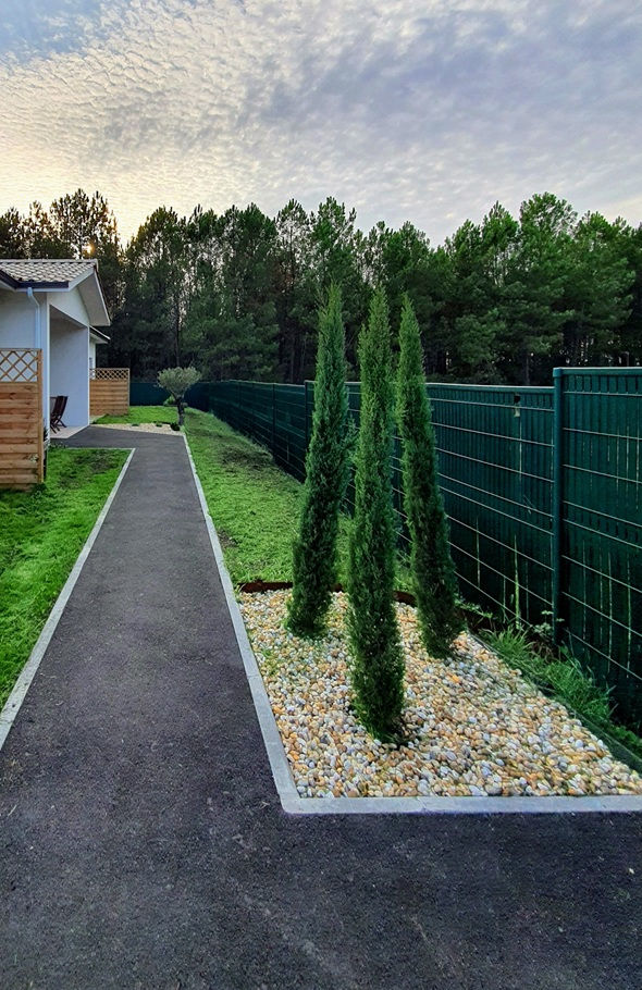 Allée bordée de cyprès et de pelouse menant à une maison moderne, entourée d'une clôture verte, avec une forêt en toile de fond sous un ciel nuageux.