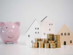 Pink piggy bank, stacks of coins, and three wooden house models on a white surface, conveying savings and investment in real estate.