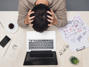 Person with head in hands over laptop, surrounded by sketches, phone, and coffee. Workspace suggests stress or frustration.