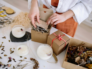 Person packing a gift box with straw and a card. Candles, dried flowers, and leaves on the white table. Red ribbon adds a festive touch.
