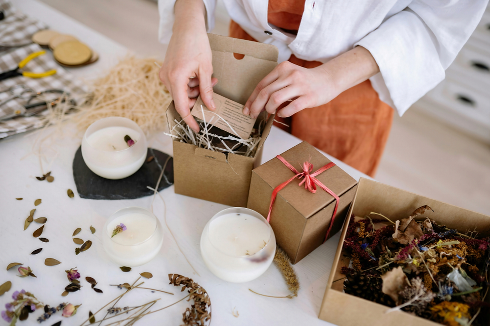 Person packing a gift box with straw and a card. Candles, dried flowers, and leaves on the white table. Red ribbon adds a festive touch.