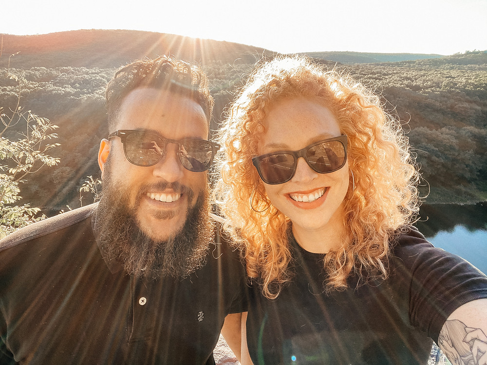 Black man and white woman smiling into camera on a mountaintop wearing sunglasses