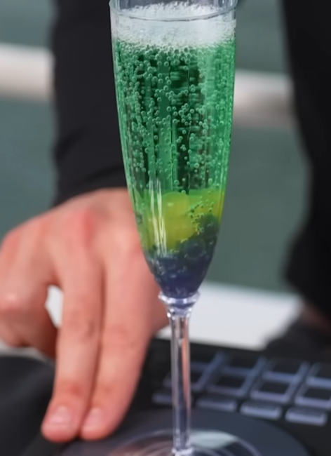 A tall champagne flute containing a carbonated green beverage with visible layers of yellow and dark blue boba pearls at the bottom, creating a gradient effect.