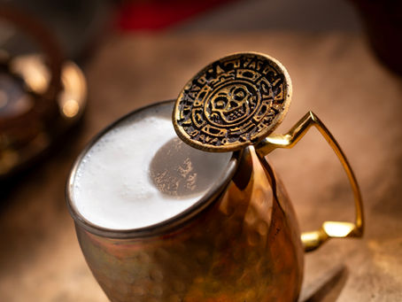 A bronze cup with frothy liquid is topped by a decorative metal disc with intricate designs. Warm lighting creates a cozy ambiance.