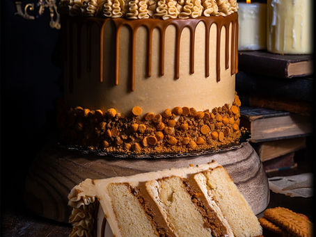 Layered cake with cookie butter drip and frosting rosettes sits on a wooden stand. A slice is cut showing the vanilla cake, cookie butter filling and butterscotch buttercream. Candles, a chandelier and books show in the dim background.