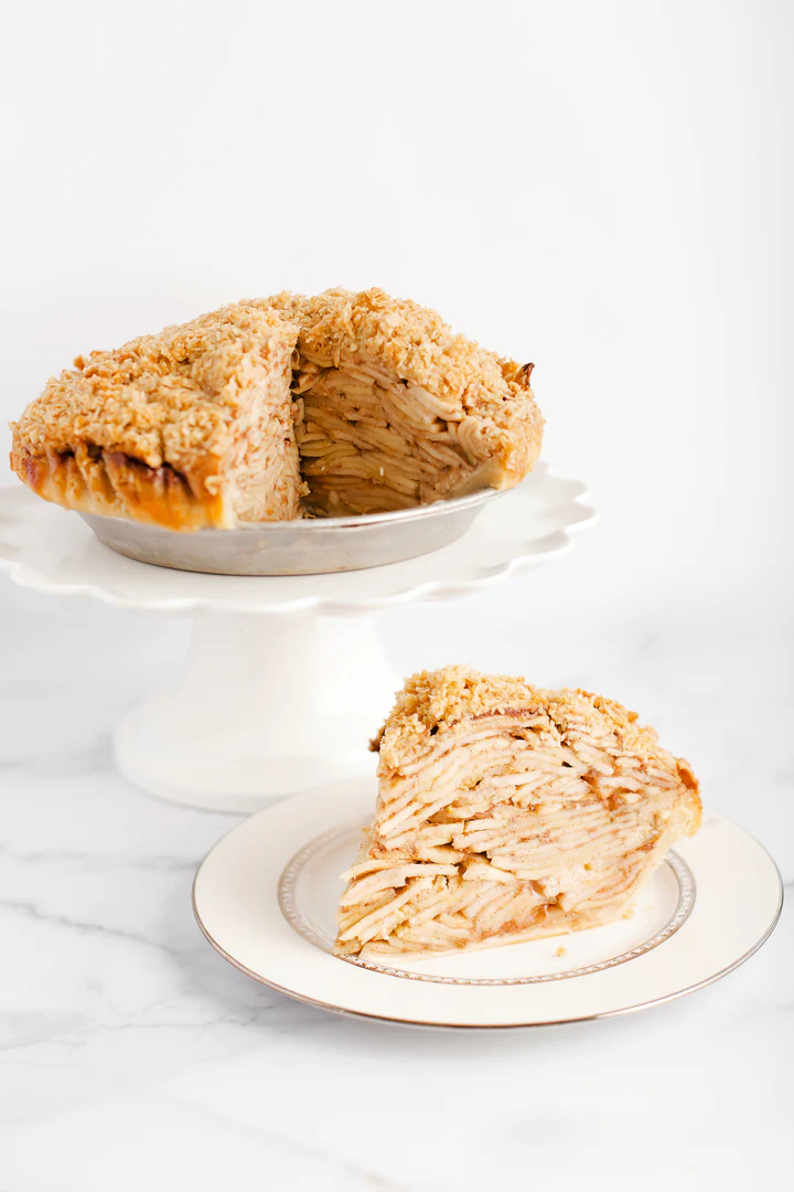 A tall deep-dish apple pie with a golden crumb topping, filled with tightly layered apple slices, displayed on a white cake stand with a single slice served on a decorative plate in the foreground. It is the Apple Crumble Pie at Cake Bake Shoppe