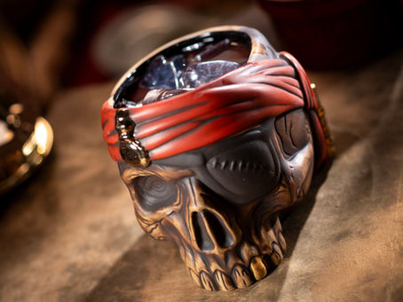 A close-up of a pirate-themed skull mug from The Beak and Barrel at Magic Kingdom, filled with an iced beverage. The mug features intricate details including a stitched eye patch, golden tooth, and a red bandana with gold accents, evoking a swashbuckling, adventurous feel.