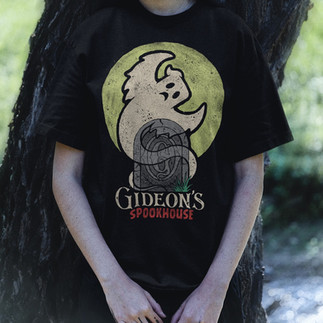 Person wearing a black “Gideon’s Spookhouse” T-shirt featuring a ghost and tombstone logo with yellow moon backdrop, standing against a tree in a shadowy outdoor setting.