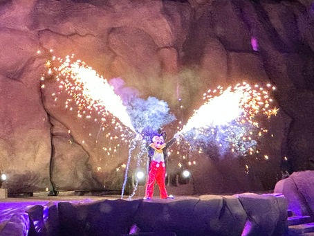 Mickey Mouse performing in the grand finale of "Fantasmic!" at Disney's Hollywood Studios. Dressed in his classic red pants and a tuxedo jacket, Mickey stands atop a rocky mountain peak as vibrant white and orange sparks erupt from his hands. The scene is illuminated by dramatic purple and pink spotlights against a dark, rocky background.