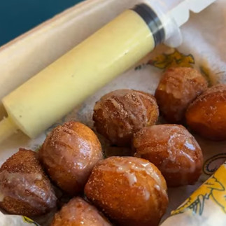 A tray of golden-brown sweet garam masala donuts drizzled with glaze sits beside a large syringe filled with creamy sauce, ready for filling or dipping. The donuts look warm, fluffy, and dusted with sugar, creating a playful and indulgent presentation.