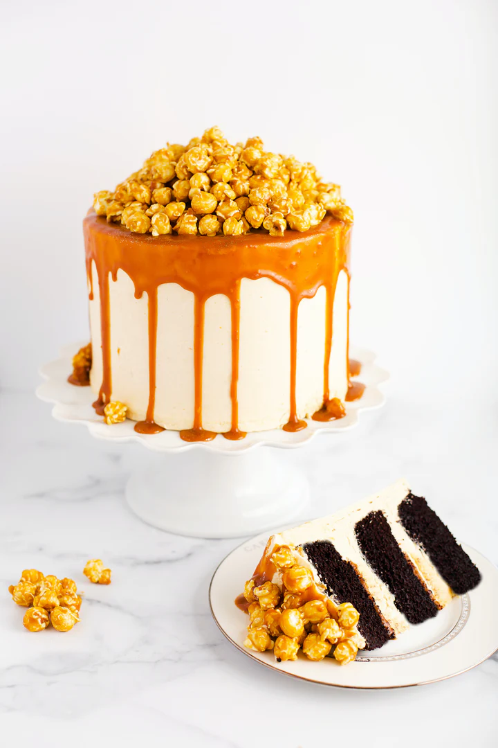 A tall chocolate layer cake covered in smooth caramel-drizzled frosting, topped with a generous pile of caramel popcorn, displayed on a white cake stand with a slice cut out and plated beside it. It is the Caramel Popcorn Chocolate Cake at the Cake Bake Shoppe.