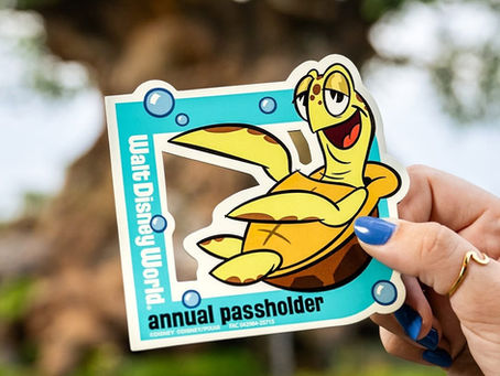A hand with blue-painted nails holds a colorful Walt Disney World Annual Passholder magnet featuring Crush the sea turtle from Pixar’s Finding Nemo. The magnet has a bright blue border with cartoon bubbles and the words "Walt Disney World" and "annual passholder" printed on it. The background shows a blurred view of the Tree of Life at Disney's Animal Kingdom.