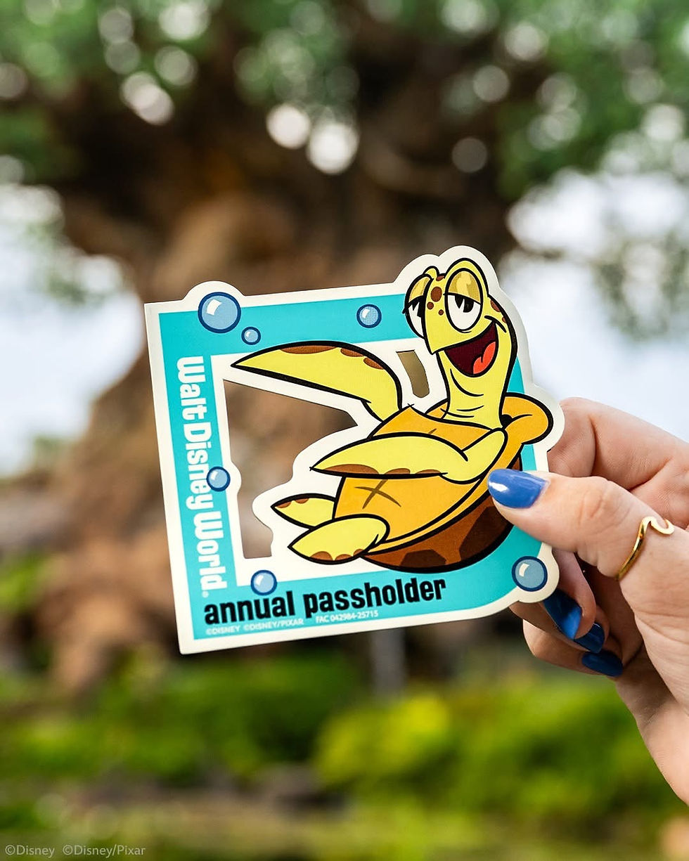 A hand with blue-painted nails holds a colorful Walt Disney World Annual Passholder magnet featuring Crush the sea turtle from Pixar’s Finding Nemo. The magnet has a bright blue border with cartoon bubbles and the words "Walt Disney World" and "annual passholder" printed on it. The background shows a blurred view of the Tree of Life at Disney's Animal Kingdom.