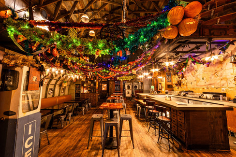 A warmly lit bar decorated for Halloween with colorful garlands, tinsel, and pumpkin lanterns hanging from the wooden ceiling. The rustic interior features long wooden tables with metal stools, a bar lined with glasses and bottles, and cozy booth seating along the wall. The festive decorations include glowing lights, spooky cutouts, and hanging jack-o’-lanterns, creating a vibrant and playful seasonal atmosphere. The Interior of Jock Lindsey's Decorated for Halloween