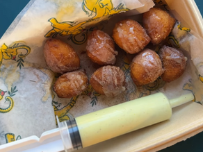 A tray of golden-brown sweet garam masala donuts drizzled with glaze sits beside a large syringe filled with creamy sauce, ready for filling or dipping. The donuts look warm, fluffy, and dusted with sugar, creating a playful and indulgent presentation.