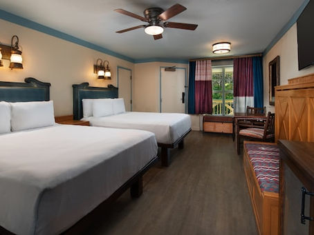Hotel room with two queen beds, wooden furnishings, overhead fan, and a window with red and blue curtains. Warm lighting creates a cozy feel. This is a guest room at Disney's Port Orleans Riverside Resort.