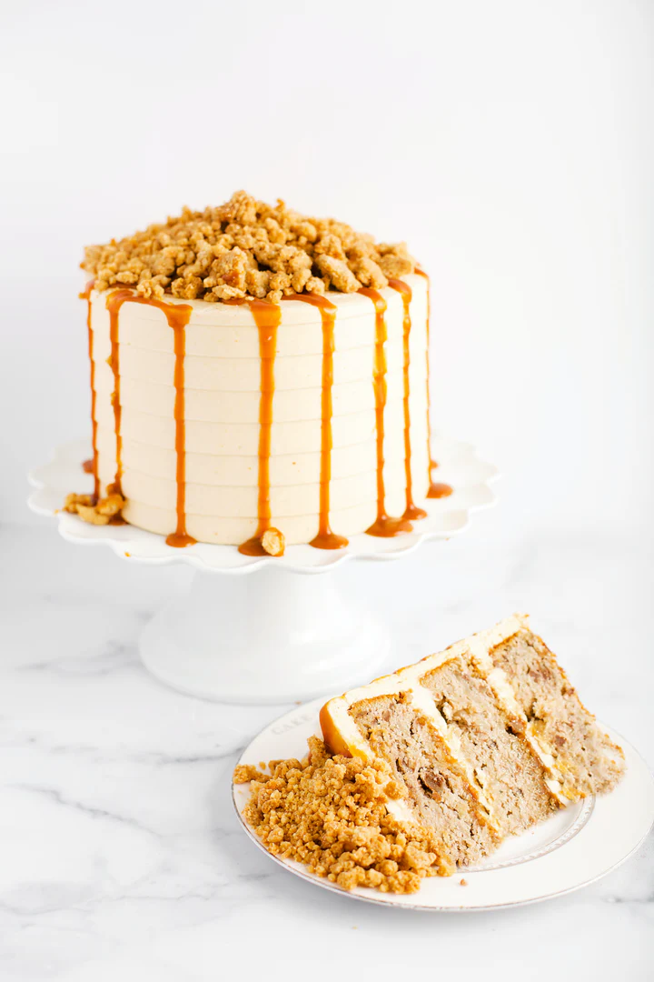A tall apple crumble cake frosted in smooth cream-colored icing with caramel drizzle down the sides, topped with a layer of crumbly streusel, displayed on a white cake stand with a slice served on a plate, showing moist apple cake layers with crumble topping scattered around. It is the Apple Crumble Cake at Cake Bake Shoppe
