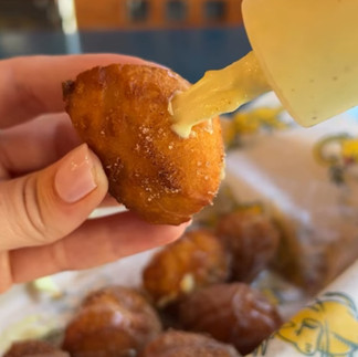 A hand holds a sweet garam masala donut while filling it with creamy sauce from a large syringe. More golden-brown donuts sit in a tray below. This is a close up view of the Sweet Garam Masala Donut from eet