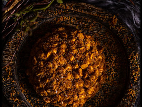 Close-up of a round, crumbly cookie with butterscotch chips and cookie cumbles on top. The cookie is on a dark, textured plate. Surrounding twigs and dark background create a cozy, rustic mood.