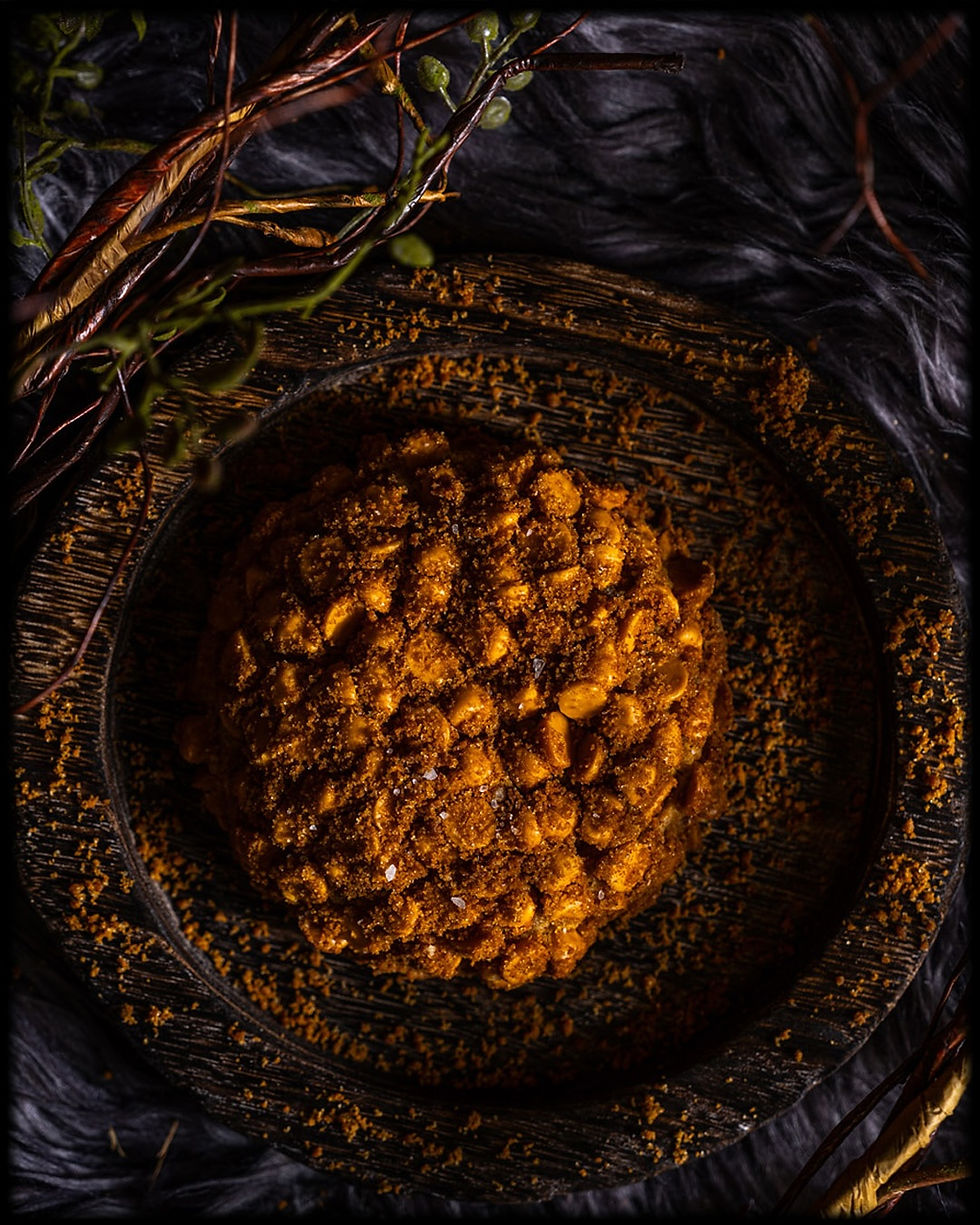 Close-up of a round, crumbly cookie with butterscotch chips and cookie cumbles on top. The cookie is on a dark, textured plate. Surrounding twigs and dark background create a cozy, rustic mood.