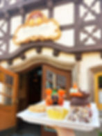 A tray of Halloween-themed caramel treats held up in front of the Karamell-Küche shop at EPCOT’s Germany Pavilion. The tray features a Mickey pumpkin caramel apple, a Minnie witch hat caramel apple, a small caramel tart, and a chocolate-covered caramel treat drizzled with white icing.