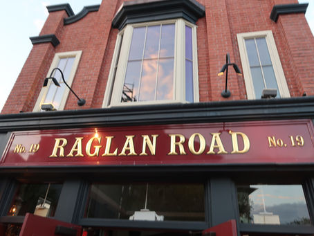Red brick building facade with large window reflects sky. Sign reads "Raglan Road No. 19" in gold letters. Warm, inviting atmosphere.