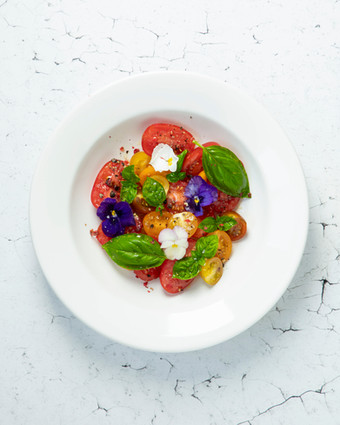 Frischer Tomatensalat mit Basilikum und violetten essbaren Blüten auf weißem Teller, ideal für gesunde Food-Fotografie und gastronomische Präsentationen.