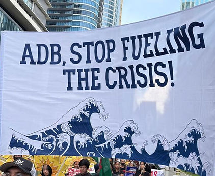 Protesters hold a white banner reading ADB, STOP FUELING THE CRISIS! with blue wave art in a city street.