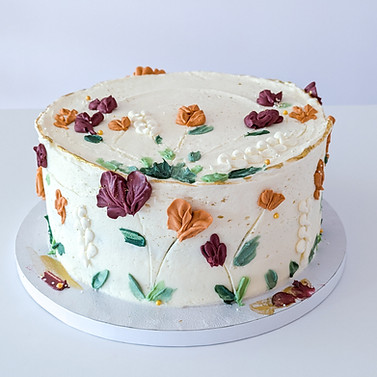 Photo of a cake with burnt orange and dark red florals piped on sides and top of cake, with gold accents and various shades of greenery