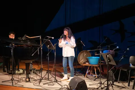 Fotografia de uma mulher a cantar com um homem a acompanhar ao piano.