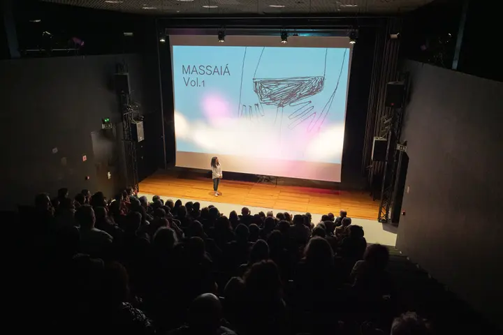 Fotografia de um auditório com plateia cheia em silhueta. No palco está uma mulher de pé com uma projeção de fundo de um desenho simples do corpo de uma mulher e escrito "Massaiá Vol1".