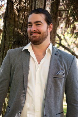 Retrato de um fotográfico de um rapaz sorridente. Tem um rabo de cabalo, barba aparada e tem um fato cinzento e uma camisa branca.