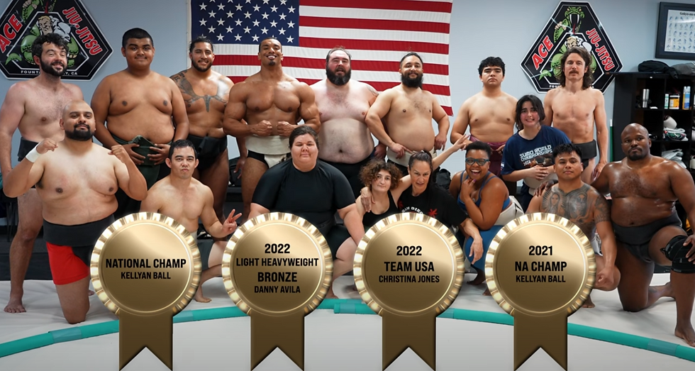 A group of sumo wrestlers pose smiling in a gym, with an American flag backdrop. Medals list achievements like National Champ Kellyan Ball.