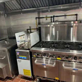 Close-up of the gas line installation for two commercial countertop burners, showing black pipes labeled with yellow "NATURAL GAS" markers, valves, and a gas regulator.