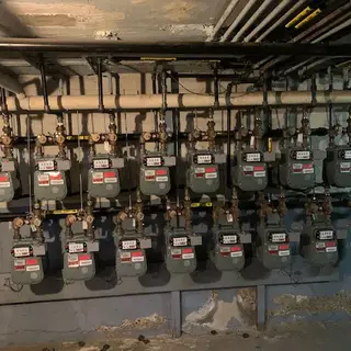 A row of multiple residential or commercial gas meters mounted on a wall with extensive piping and valves.