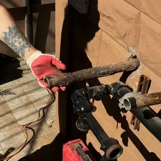 A gloved hand holding a severely rusted, cut section of pipe near a new black plumbing connection on a textured brown wall.