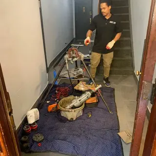 A plumber working on a pipe assembly on the ground in an indoor utility space, surrounded by large pipes, tools, and an oil pan.