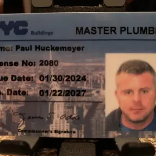 Close-up of an NYC Buildings Master Plumber license ID card for Paul Huckemeyer, showing his photo, license number 2080, and issue/expiration dates against a cityscape background.